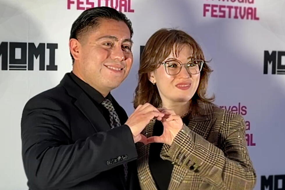 Two smiling people standing side by side form a heart shape with their hands at a festival photo backdrop, showing camaraderie and celebration.