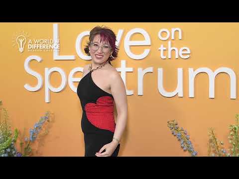 Woman in a black dress with a red heart design posing in front of an orange backdrop reading 'Love on the Spectrum'.