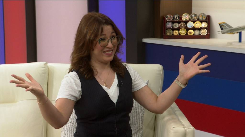 Smiling woman with glasses sits on a white sofa, arms open in a TV studio gesture.