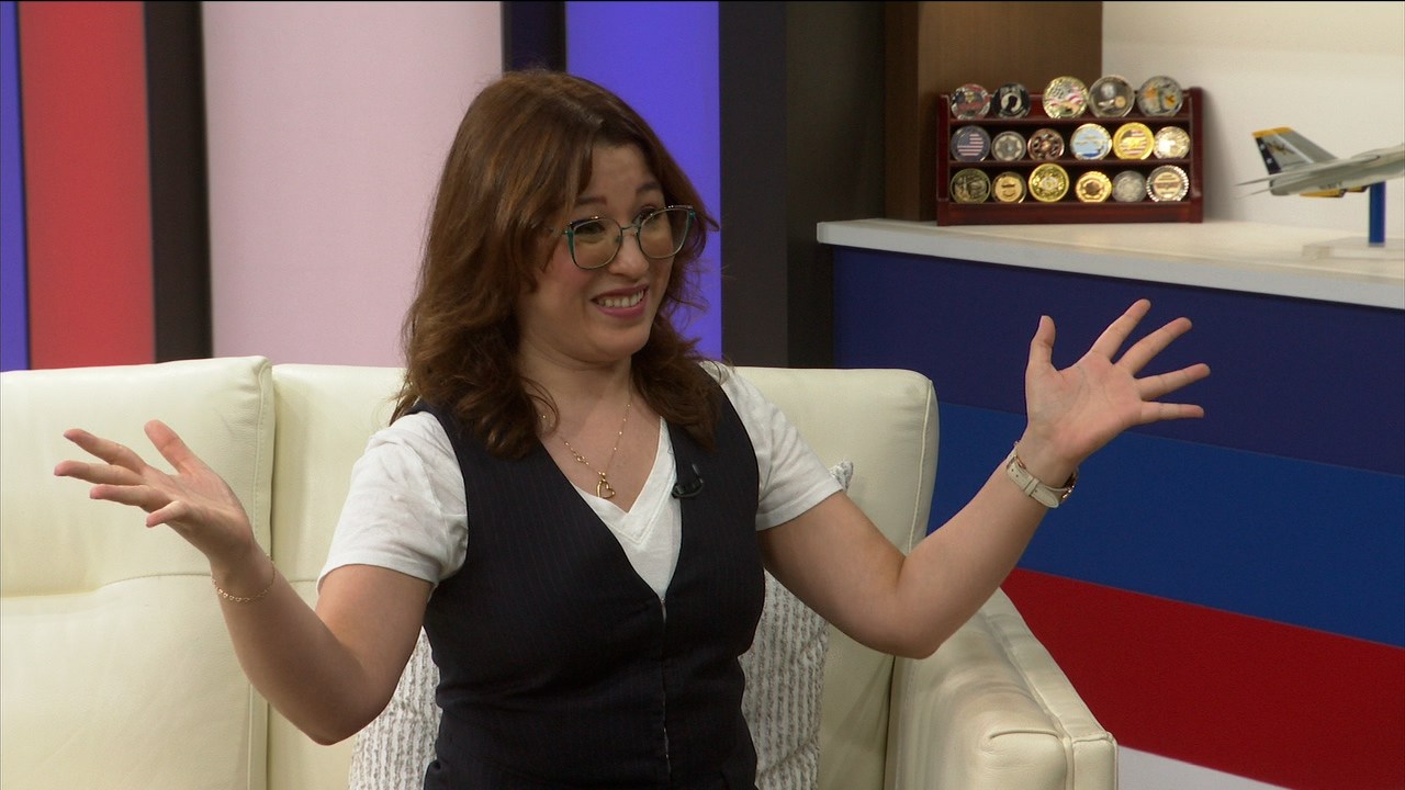 Smiling woman with glasses sits on a white sofa, arms open in a TV studio gesture.