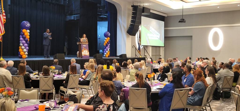 Speaker at a podium on stage presenting to a large audience; purple and orange balloon columns flank the stage at a banquet-style event.