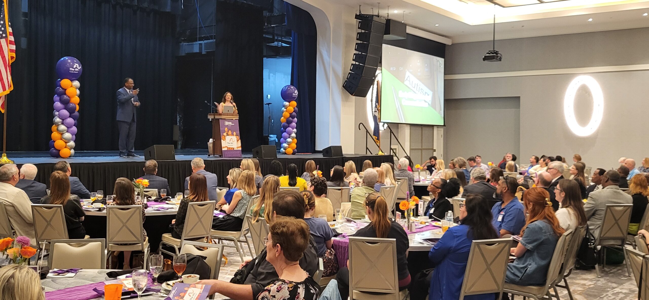 Speaker at a podium on stage presenting to a large audience; purple and orange balloon columns flank the stage at a banquet-style event.