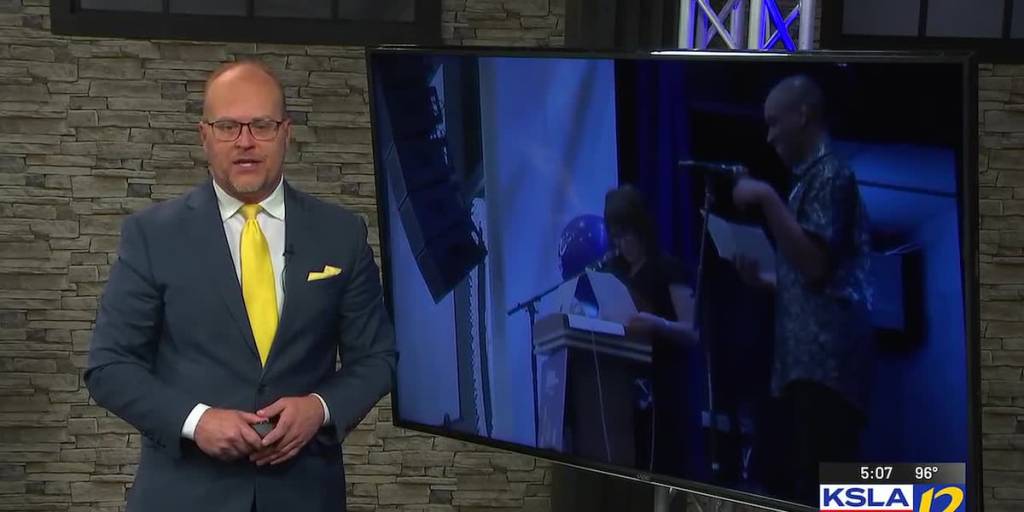 News anchor in a blue suit and yellow tie stands beside a large screen showing people at a podium on a TV set, with a stone wall backdrop.